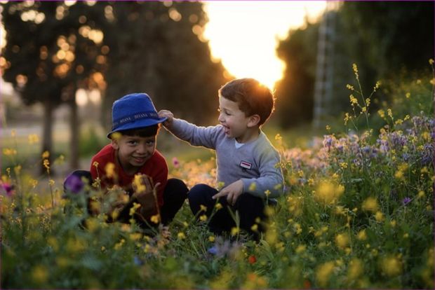 Dos niños riendo, agachados entre flores silvestres a contraluz del atardecer
